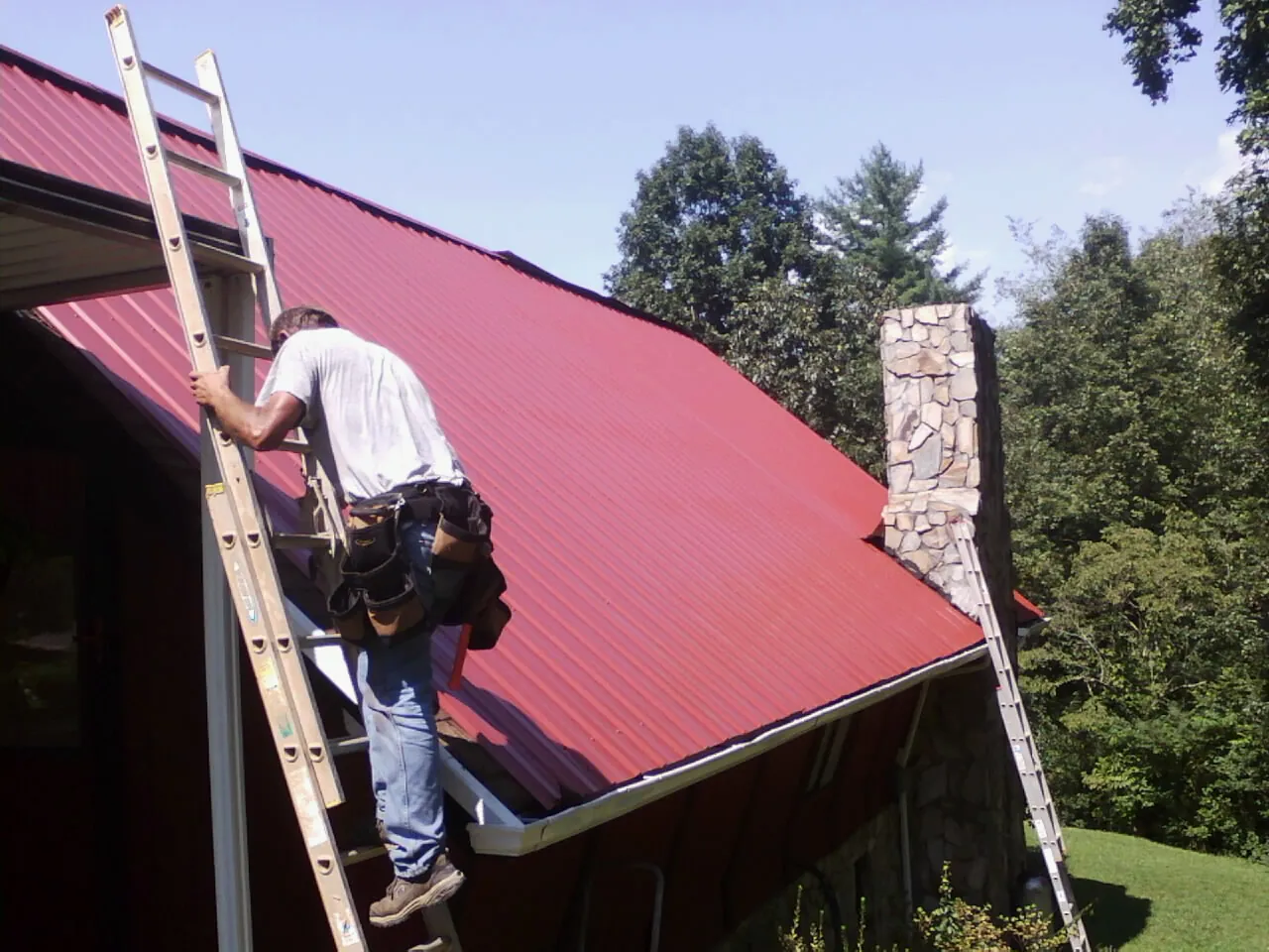 Roofer on a ladder beside a red metal roof and stone chimney — Soffit & Fascia Repair by Roofing Brothers in Yukon